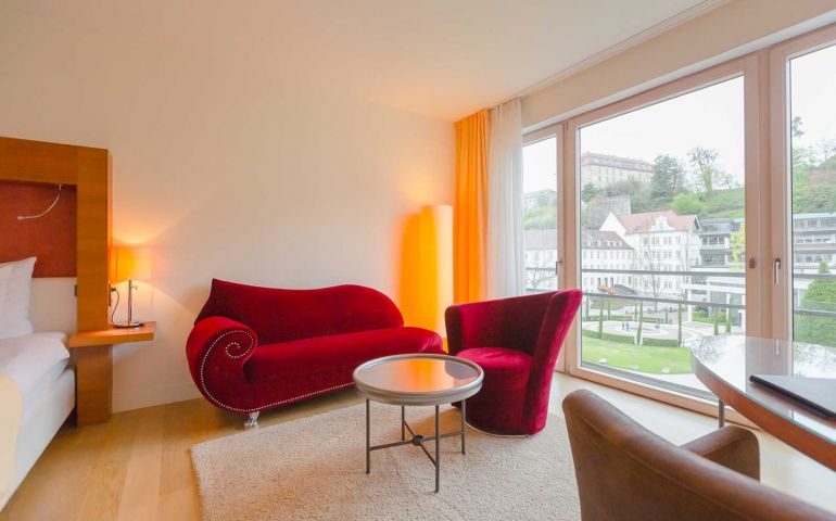 Aufnahme einer Sitzecke mit einem roten geschwungenen Sessel und einem Sofa vor einer hellen Fensterfront im Suitenhotel in Baden-Baden
