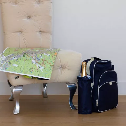 Ein blauer Rucksack mit einer Sektflasche steht neben einem beigen Stuhl auf dem eine Wanderkarte liegt
