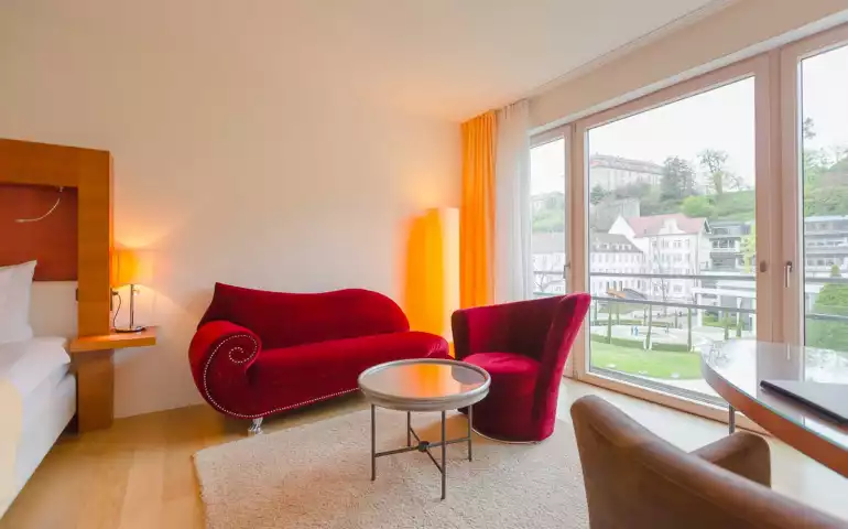Aufnahme einer Sitzecke mit einem roten geschwungenen Sessel und einem Sofa vor einer hellen Fensterfront im Suitenhotel in Baden-Baden