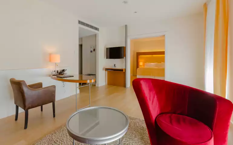 Aufnahme über einen roten geschwungenen Sessel und den Arbeitsbereich bis ins Schlafzimmer einer Suite in Suitenhotel in Baden-Baden