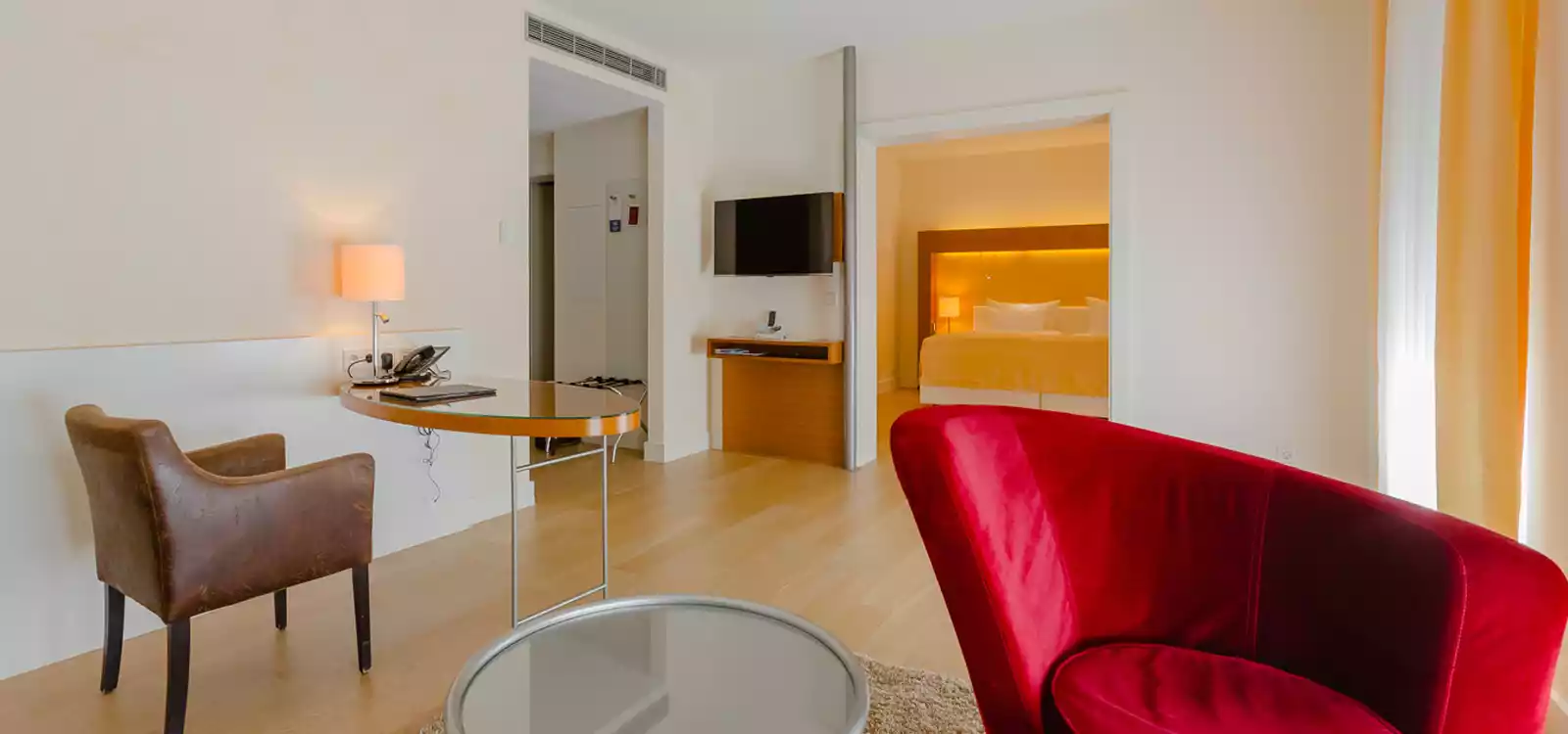 Aufnahme über einen roten geschwungenen Sessel und den Arbeitsbereich bis ins Schlafzimmer einer Suite in Suitenhotel in Baden-Baden